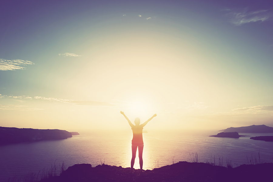 Happy woman with hands up standing on cliff over sea and islands at sunset. Vintage mood, concepts of winner, freedom, happiness etc.
